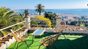 A terrace with wicker chairs and a table, overlooking a coastal cityscape.
