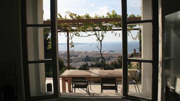 A view through a large window with a table and chairs, overlooking a cityscape and the sea.