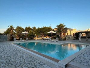 Piscina con sillas de descanso y sombrillas, rodeada de árboles y un patio de piedra.