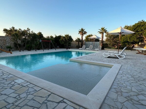 Una piscina con sillas de descanso y sombrillas, rodeada de muros de piedra y vegetación.