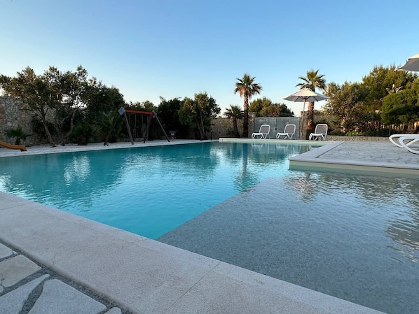 A swimming pool with clear blue water, surrounded by lounge chairs and umbrellas, and trees in the background.