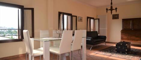 Un comedor con una mesa de vidrio y sillas blancas, un sofá negro, un mueble de madera y una alfombra estampada.