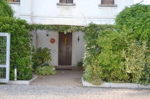 Ein Gebäudeeingang mit Holztür, einem kleinen Gartenbereich und einer weißen Wand mit einer runden Dekoration.
