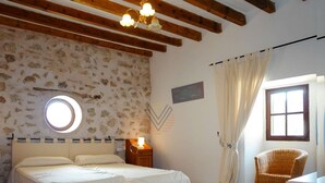 Un dormitorio con cama doble, un ventanal redondo, una mesita de noche con lámpara, una silla de mimbre y una ventana con cortinas.
