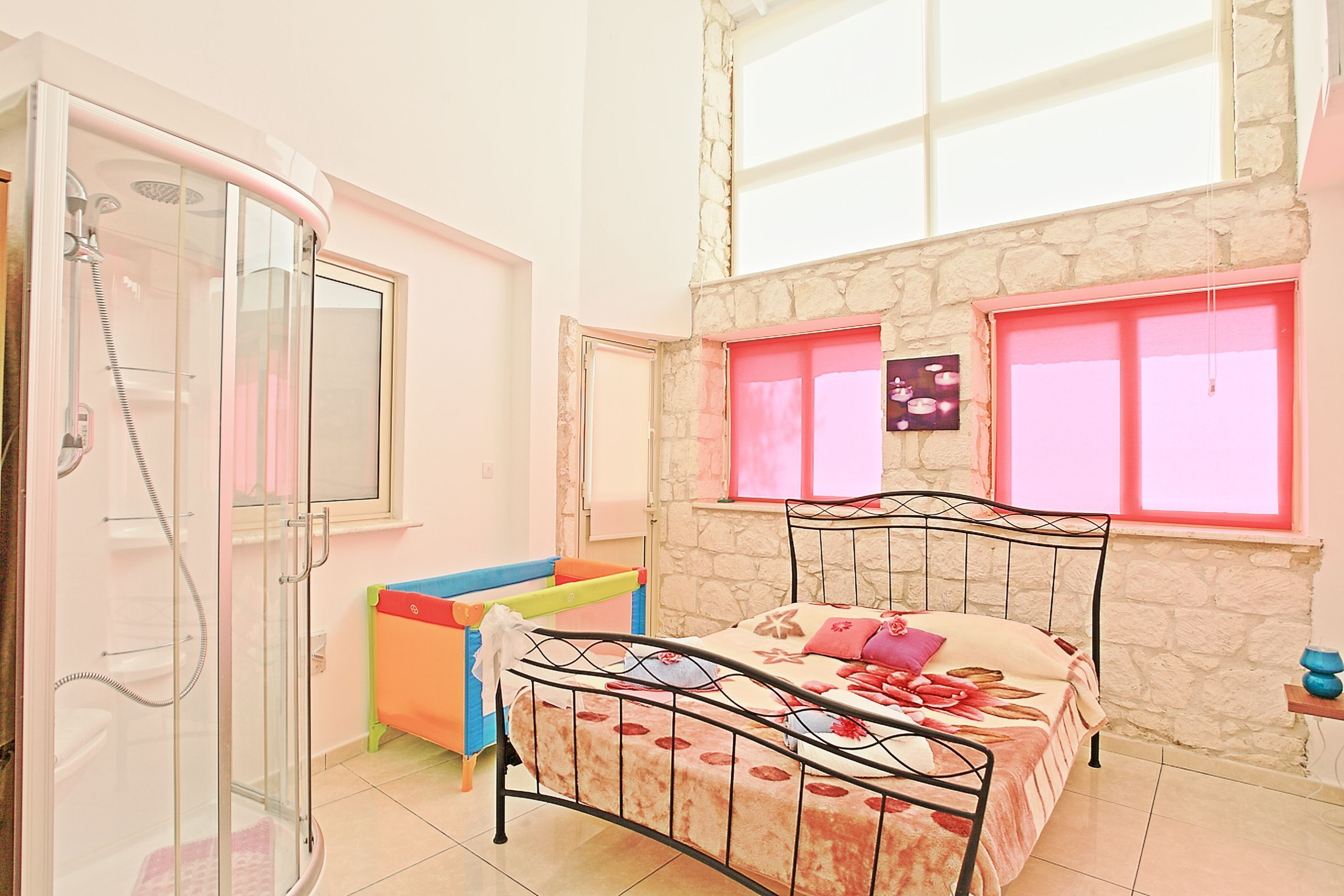 A bedroom with a bed, a shower, a window with pink curtains, and a stone wall.