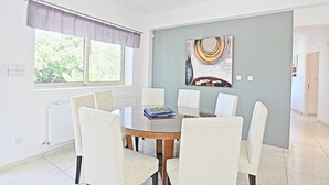 A modern dining area with a round table, white chairs, and a large window with a view of greenery. - Villa Georgios Rafaela: Secluded, pool, Wifi, A/C, sleeps 10 (Goudi Polis)