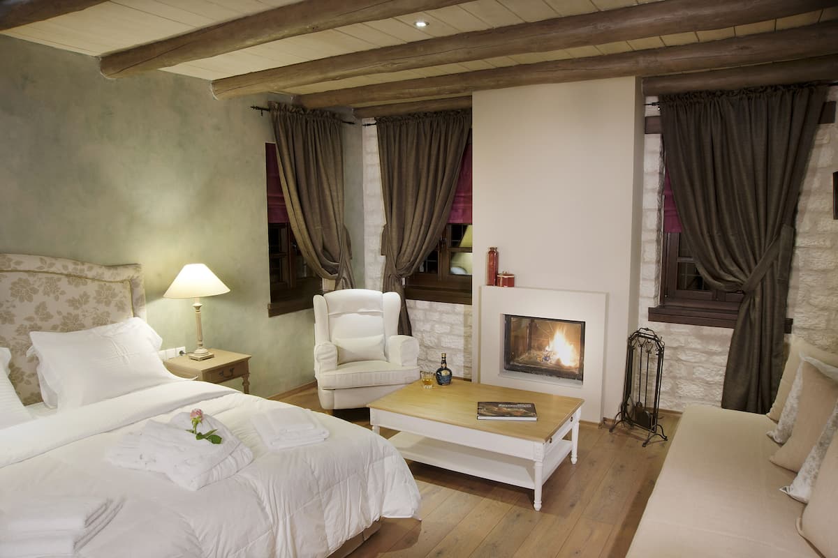 Een hotelkamer met een bed, een witte fauteuil, een houten bijzettafeltje, een open haard en gordijnen.