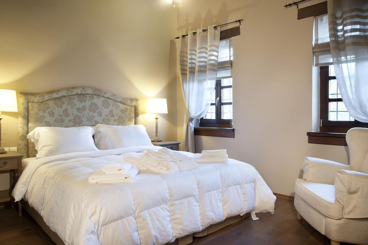 Een netjes opgemaakt bed met witte lakens en handdoeken, een hoofdbord met bloemenprint, twee bedlampjes en een raam met gordijnen.