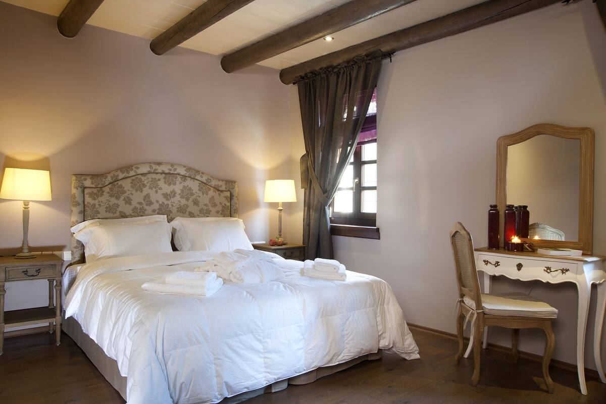 Een net opgemaakt bed met witte lakens, een houten hoofdbord, twee bedlampen, een houten stoel en een spiegel op een dressoir.