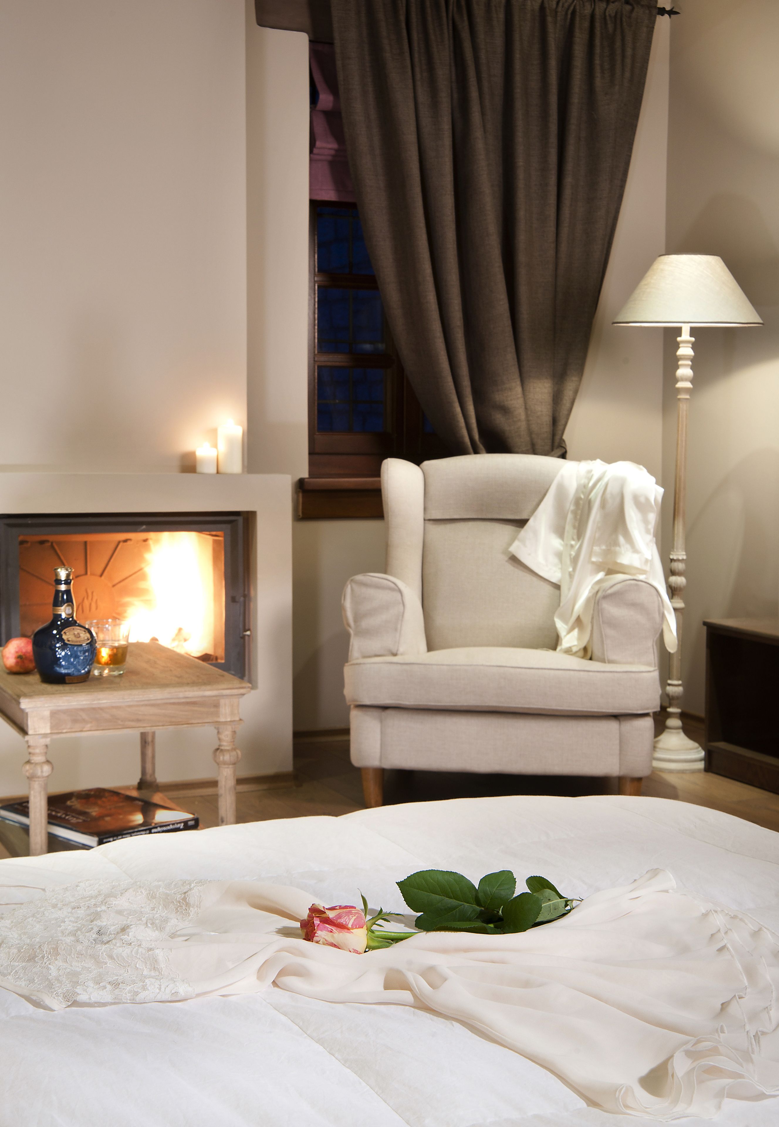 A bedroom with a fireplace, a chair, a bedside table with a bottle and glass, and a bed with a white comforter and a rose.