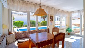 A sunlit room with a wooden table, cushioned seating, and large windows overlooking a pool and greenery. - Villa Sevilla Private Pool by Casasol (Nerja)