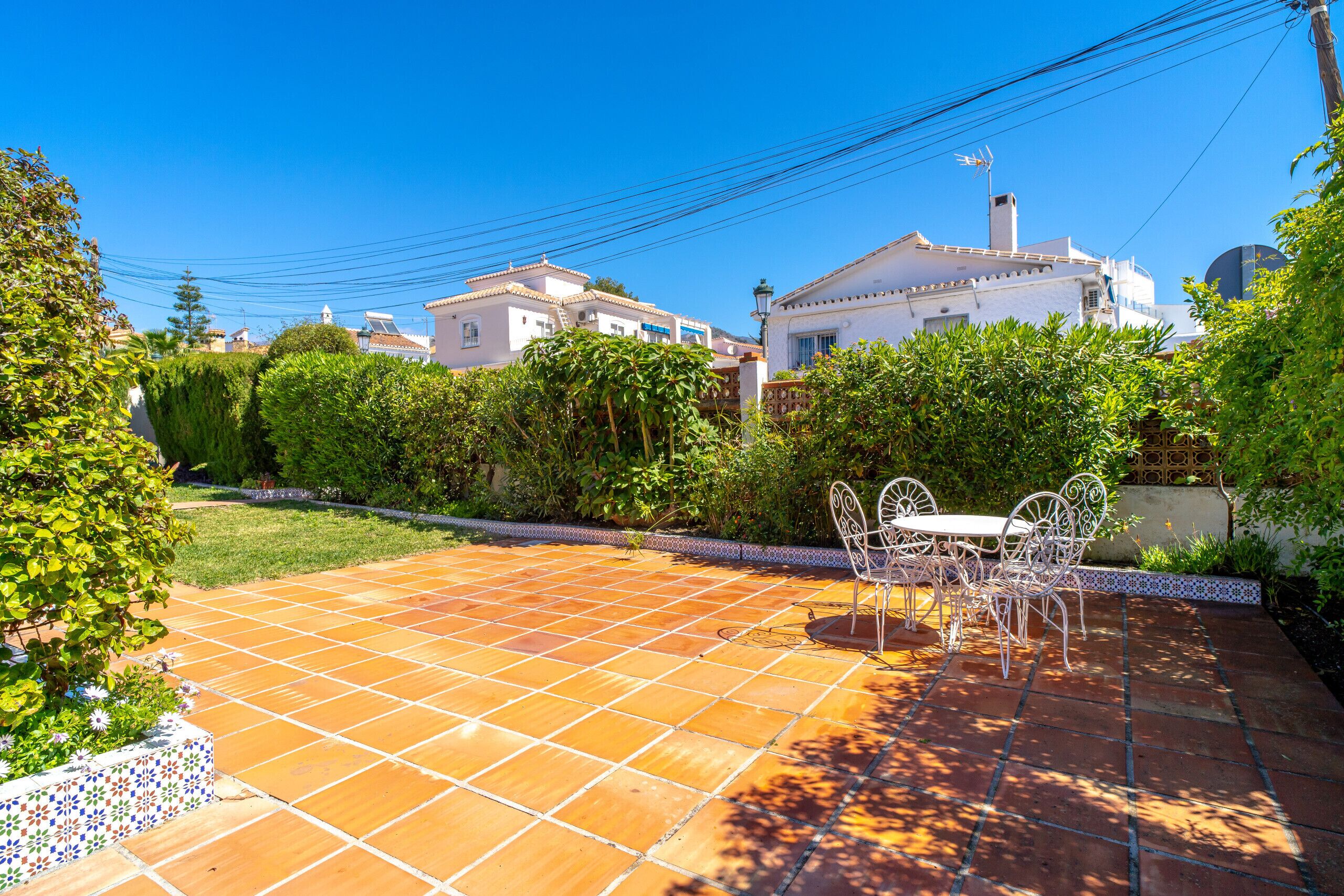 Un patio con suelo de baldosas, una mesa y sillas de metal blanco, y un jardín con diversas plantas.
