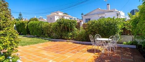 Un patio con suelo de baldosas, una mesa y sillas de metal blanco, y un jardín con diversas plantas.