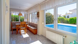 A sunlit room with a dining table, chairs, and a bench. Large windows offer views of a swimming pool and greenery. - Villa Sevilla Private Pool by Casasol (Nerja)