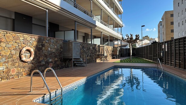Una piscina amb una paret de pedra i baranes metàl·liques, una escala de pedra i una vista d'un edifici de diversos pisos al fons.