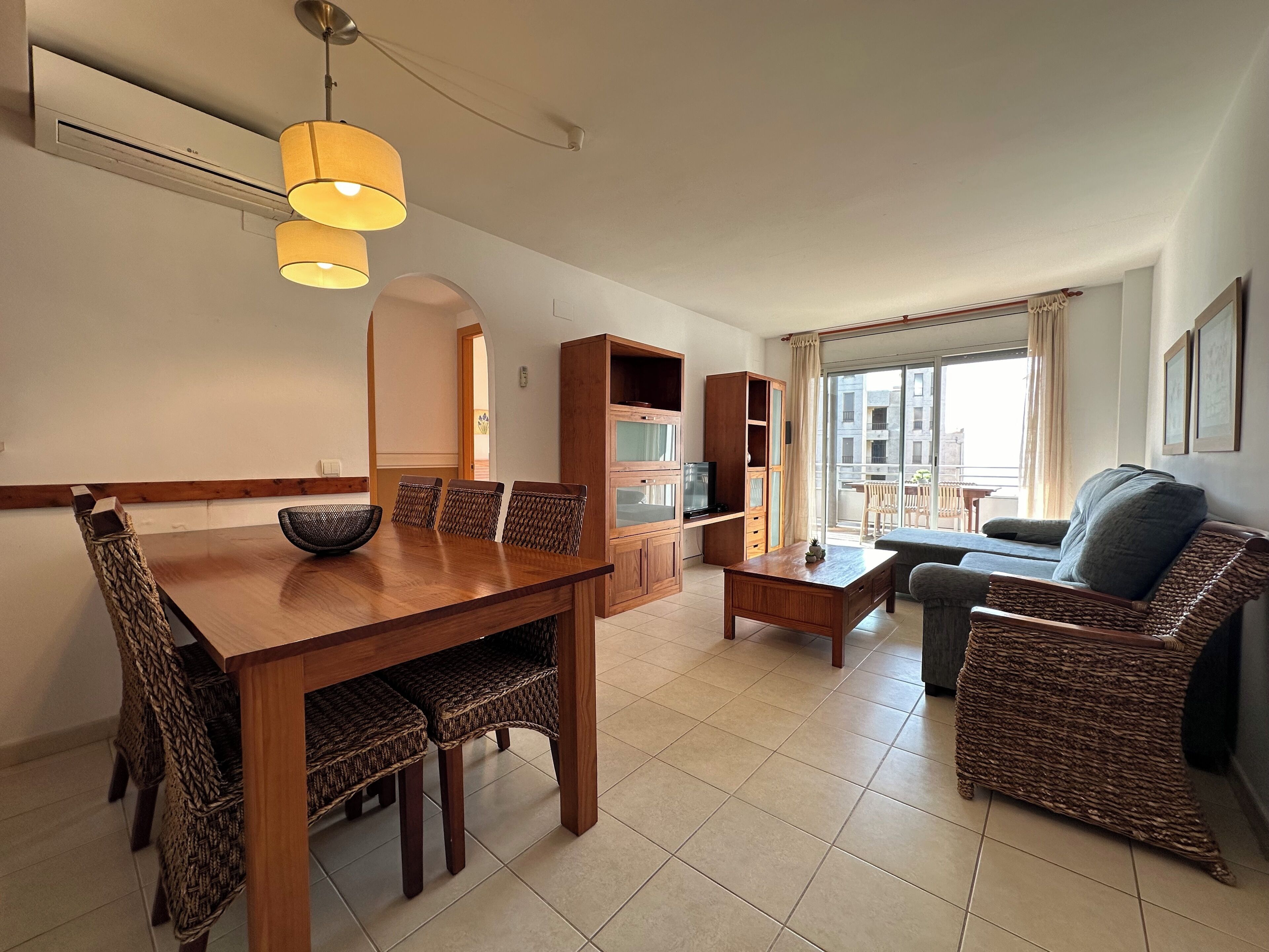 A modern living room with a dining area, wicker furniture, a TV, and a balcony with a view.