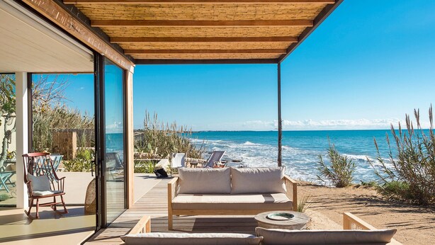 Um deck de madeira com sofá e cadeiras, com vista para uma praia e ondas.
