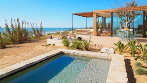 Uma área de piscina moderna ao ar livre, com águas cristalinas, rodeada por uma praia de areia e um deck de madeira com espreguiçadeiras.