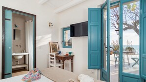 Um quarto com cama, escrivaninha, cadeira, janela com vista para o mar e banheiro com pia e espelho.