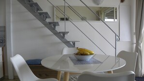 Uma área de refeições com uma mesa redonda, cadeiras brancas, uma tigela de frutas e uma escada.