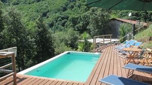 Una terraza de madera con piscina, sillas de descanso y un edificio pequeño en un área boscosa.