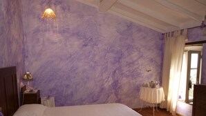 Un dormitorio con una cama grande, una mesita de noche con lámpara, una pequeña mesa con objetos decorativos y una ventana con cortinas.