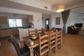 A dining area with a wooden table and chairs, a kitchen in the background, and a living room with a sofa and television.
