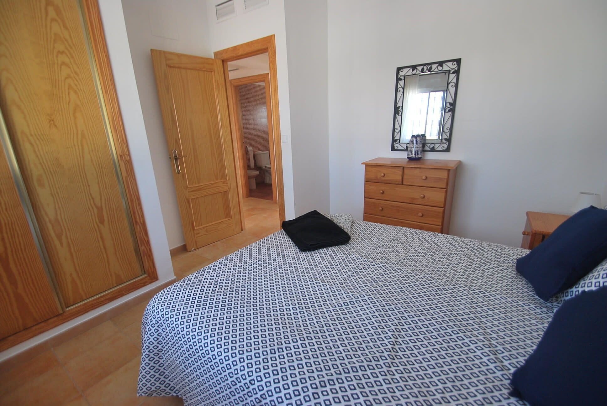 A bedroom with a patterned bedspread, a wooden dresser, a mirror, and a door leading to a bathroom.