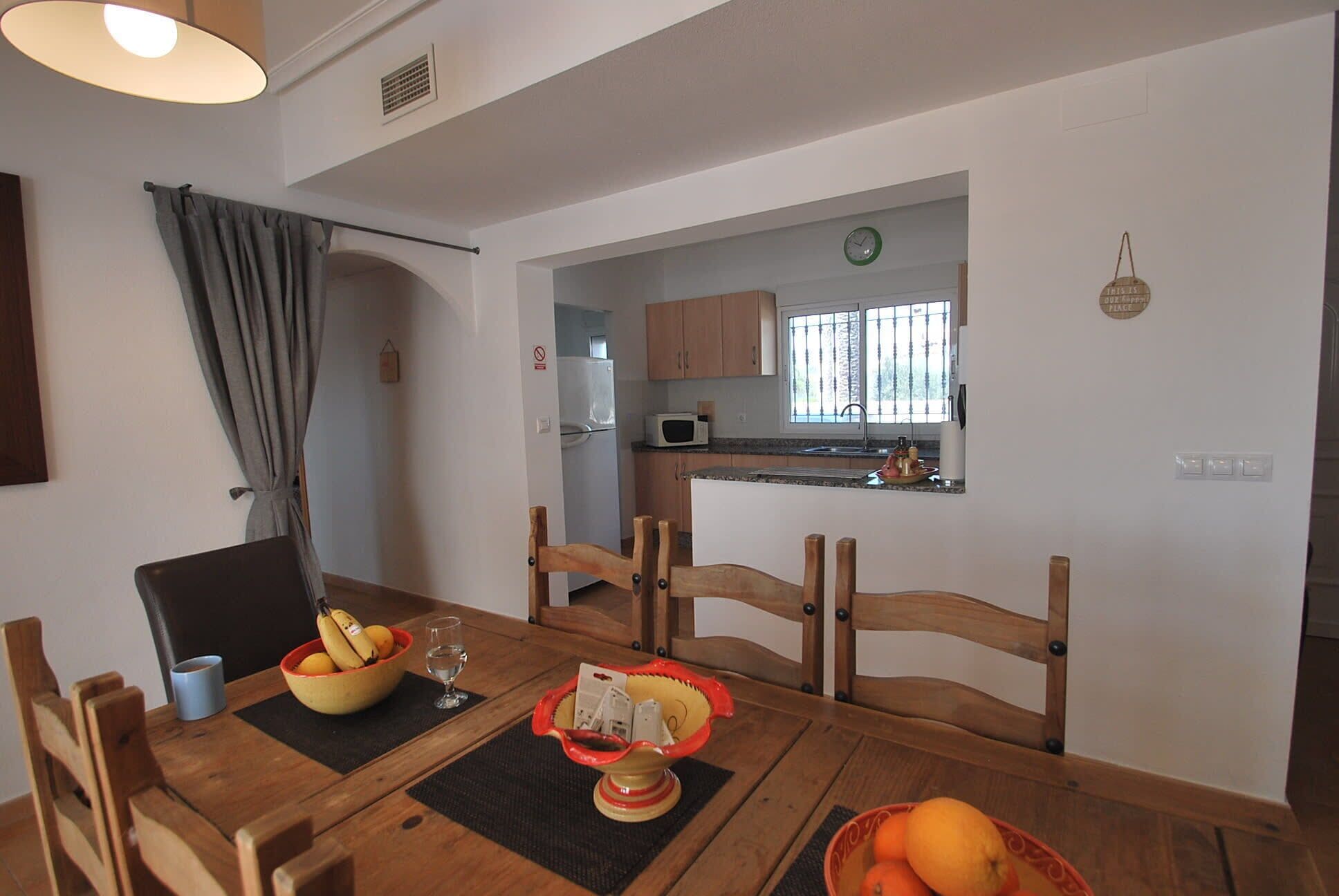A dining area with a wooden table, chairs, and a bowl of fruit. A kitchen is visible in the background with wooden cabinets and a window.