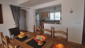 A dining area with a wooden table, chairs, and a bowl of fruit. A kitchen is visible in the background with wooden cabinets and a window. - Finca in quiet area with private pool (Catral Alicante)