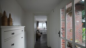 Un couloir étroit avec une commode blanche et une porte vitrée donnant sur un espace extérieur agrémenté d’un mur en briques et de verdure.