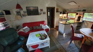 Una sala de estar acogedora con un sofá rojo, un sillón verde y una mesa de centro con un cuenco decorativo. Se ve una zona de cocina con una ventana que da a zonas verdes.