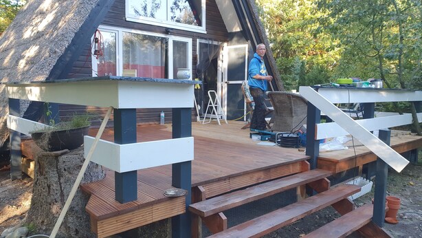 Eine Holzterrasse mit einer Bank und einem kleinen Tisch, eine Leiter und ein Haus mit geneigtem Dach.