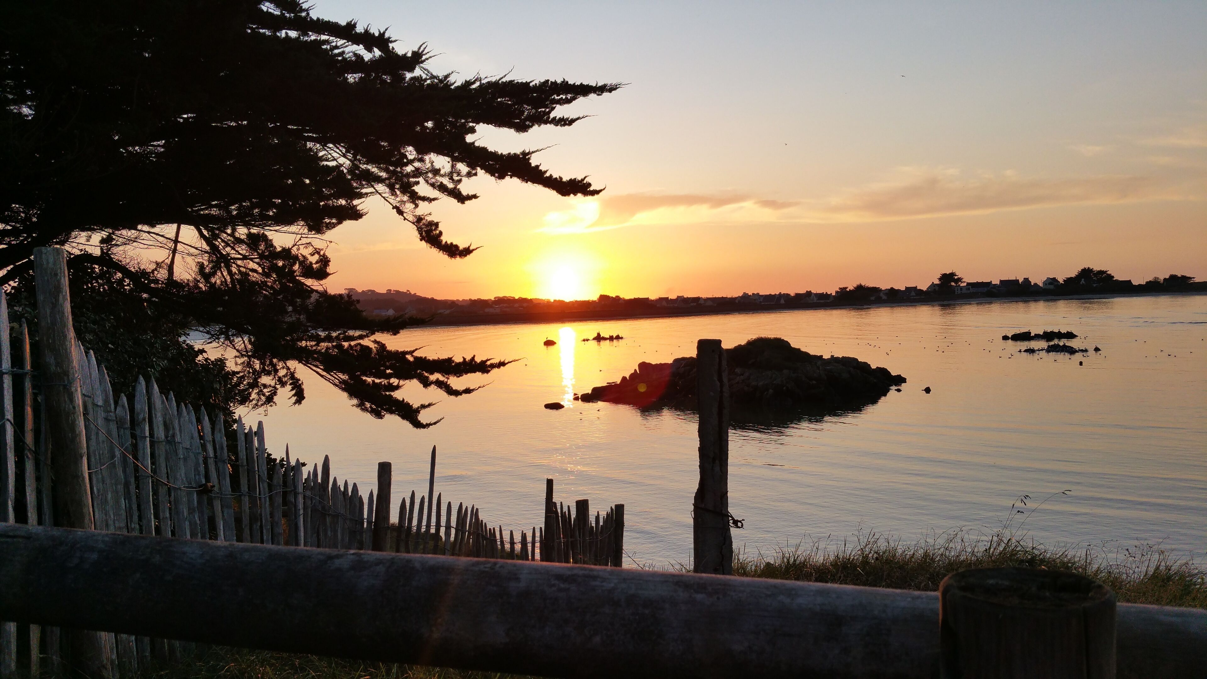 A sunset over a calm body of water with a wooden fence and trees in the foreground.