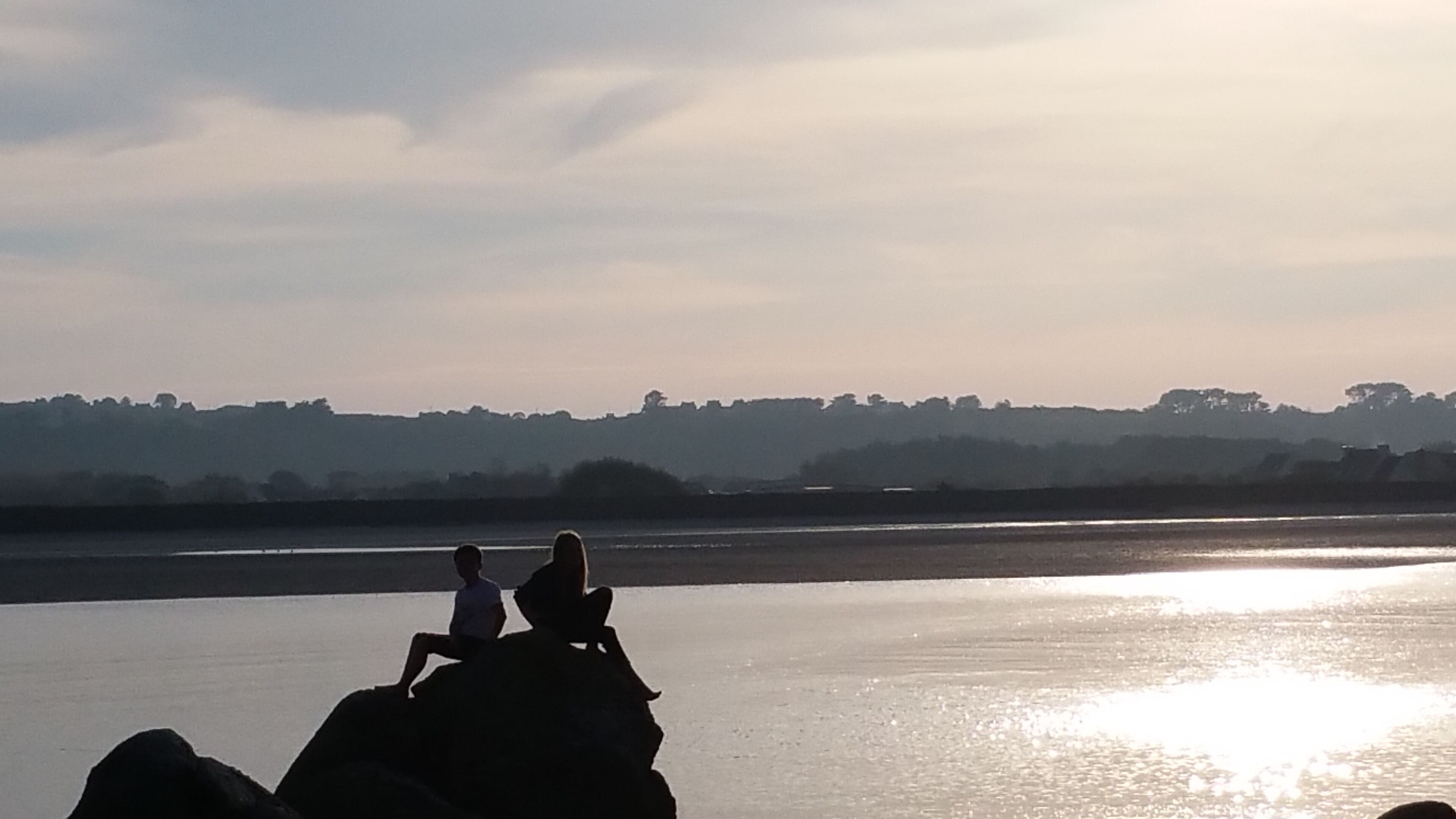 A beach scene with two people sitting on rocks. The sun is low on the horizon, creating a bright reflection on the water.