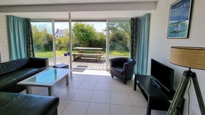 En moderne stue med sofa, sofabord, fjernsyn og lampe. Der er en skydedør af glas, der fører ud til en terrasse med et bord og stole.