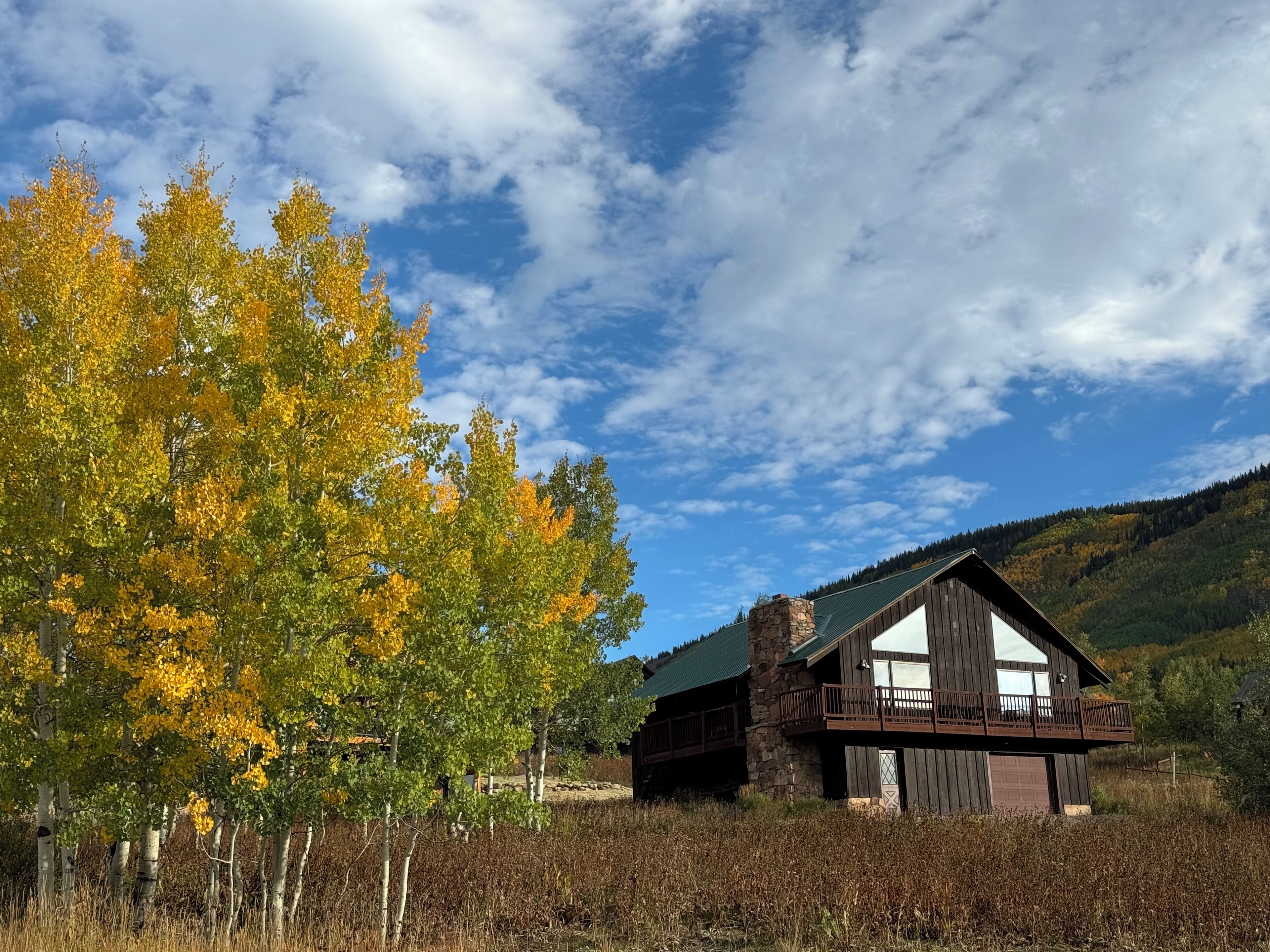 A rustic cabin with a stone chimney, wooden deck, and a view of a forested hillside.