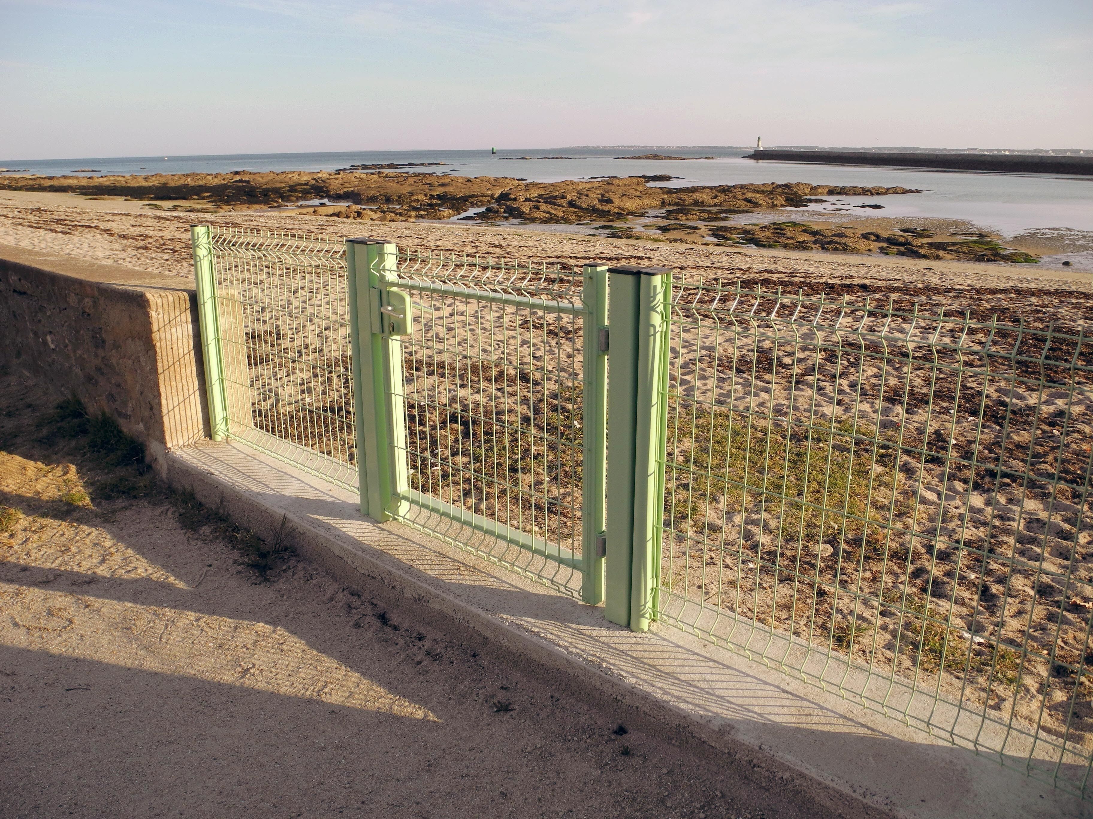 Een kustgezicht met een zandstrand, een groen metalen hek en een vuurtoren in de verte.