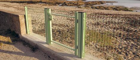 Een kustgezicht met een zandstrand, een groen metalen hek en een vuurtoren in de verte.