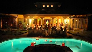 A poolside gathering with people seated around a pool, a building with arched windows, and a well-lit outdoor area.