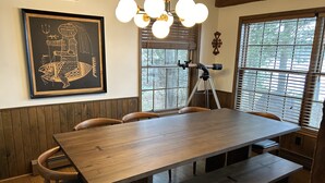 A dining area with a wooden table and benches, a framed artwork on the wall, a telescope, and windows with blinds.