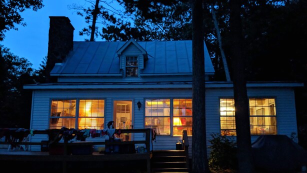 A white house with a metal roof and large windows. There is a porch with benches and a pathway leading to the house.