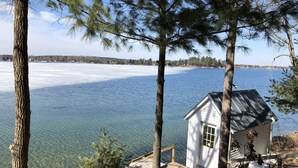 A small white cabin with a metal roof, surrounded by trees and overlooking a frozen lake.