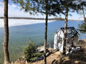 A small white cabin with a metal roof, surrounded by trees and overlooking a frozen lake.