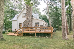 Une maison avec une cheminée en pierre, une terrasse en bois et un espace salon extérieur entouré d’arbres.