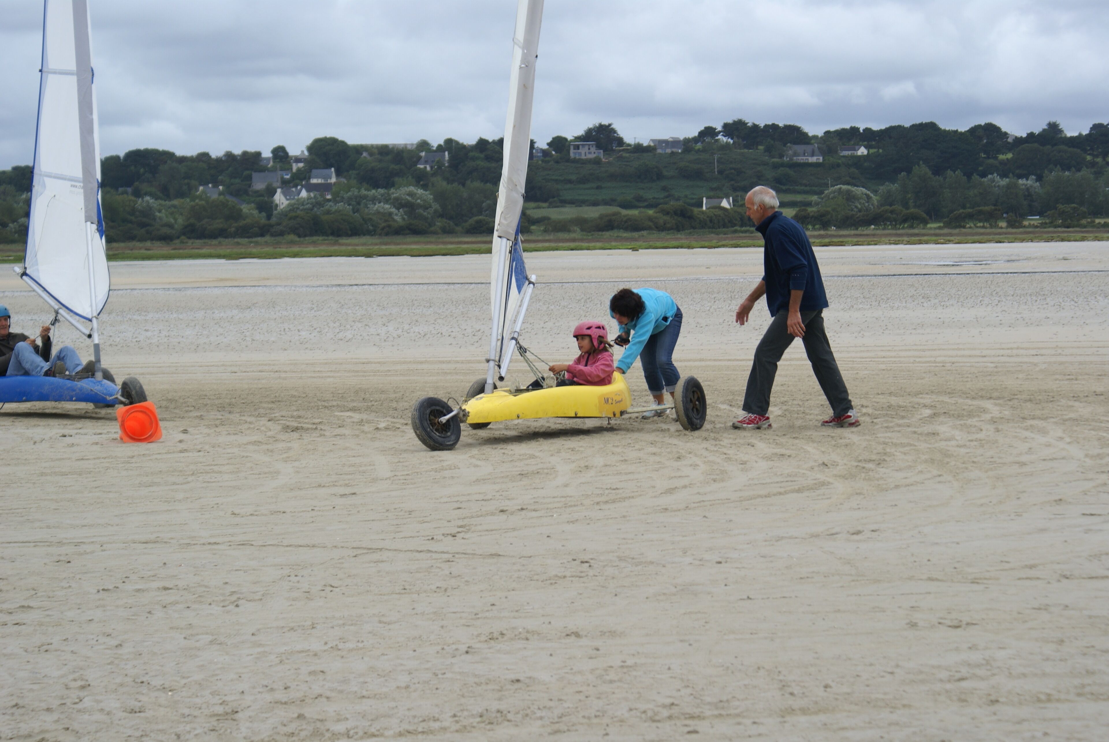 Une plage de sable avec un voilier et une personne aidant un enfant à bord d’un petit voilier.