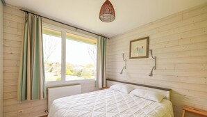 A bedroom with a bed, wooden walls, a window with curtains, a radiator, and a hanging light fixture.