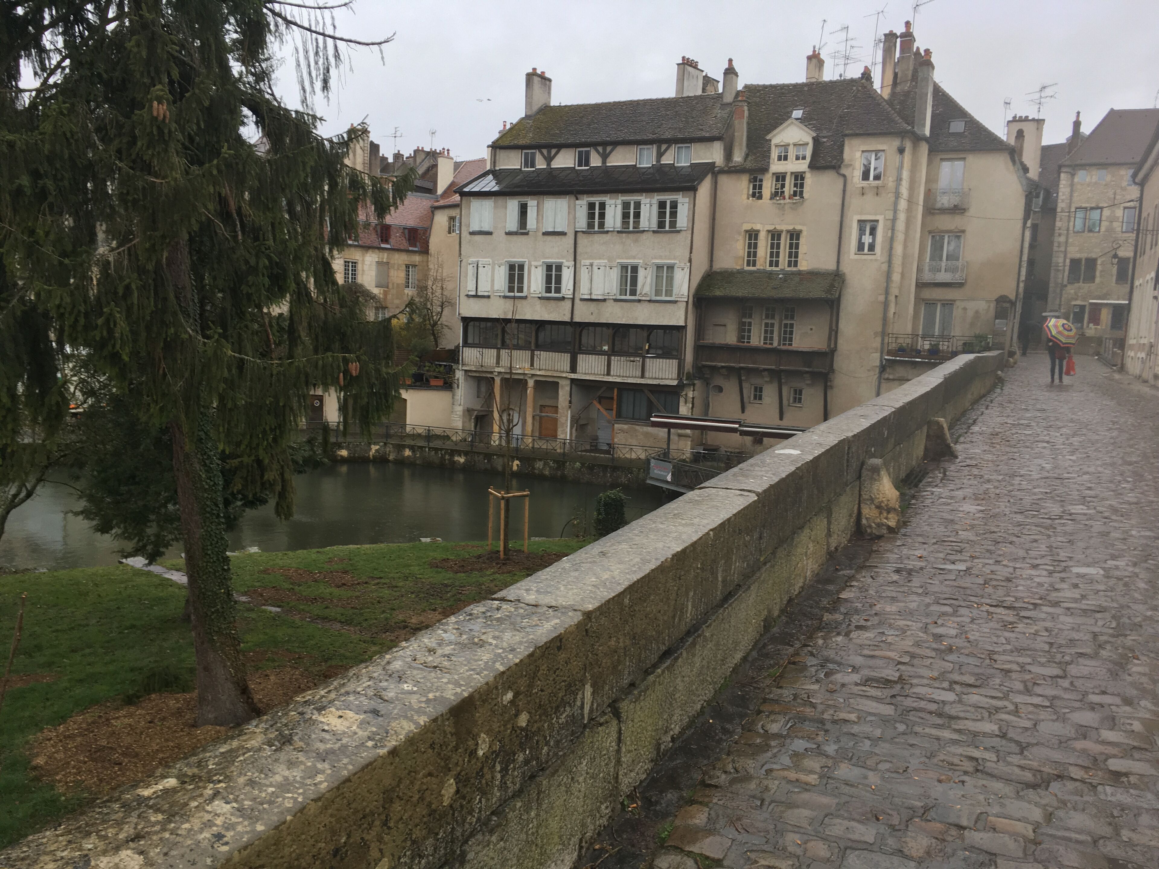 Un sentier pavé de pierres, longeant un mur de pierre d’époque, typique de l’architecture européenne traditionnelle, avec un plan d’eau et un arbre.