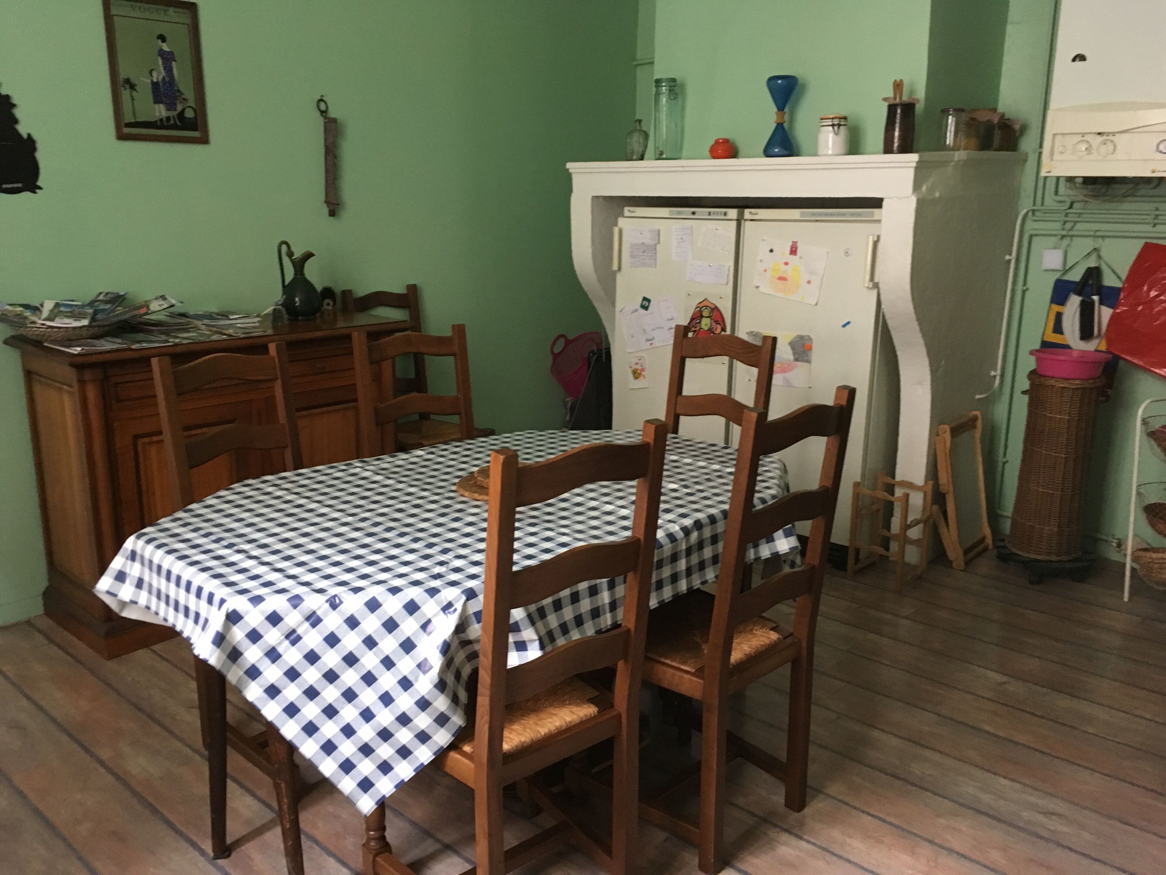 Un coin repas avec une nappe à carreaux, des chaises en bois, un réfrigérateur recouvert de papiers et un mur vert orné d’un tableau encadré.