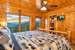A wooden cabin with a bed, a ceiling fan, a TV, and a balcony with a mountain view.
